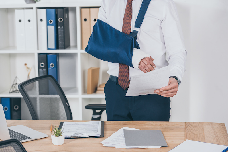 Worker with broken arm in bandage standing and holding paper over table in office, compensation